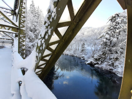 Snow-covered steel truss bridge over a dark river surrounded by snowy forested hills.