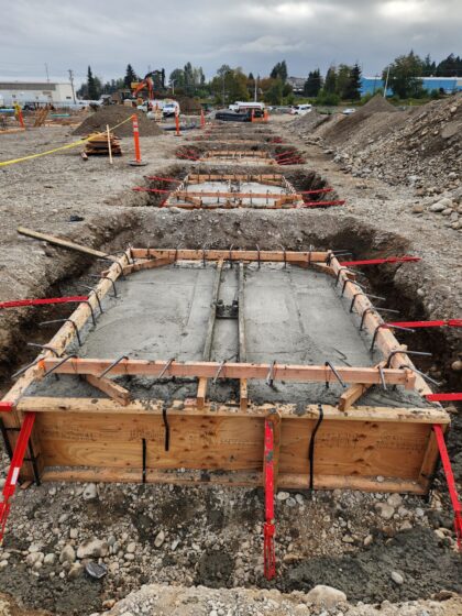 Construction site showing a freshly poured concrete foundation with wooden forms and exposed rebar, excavated trenches, machinery, and soil piles.