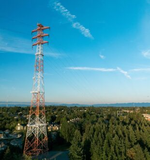 Tacoma Public Utilities Potlatch line tower in Tacoma's west slope community. The red and white transmission tower is shown from a drone on a sunny late afternoon with a blue sky and wispy clouds in the background. Evergreen trees surround the tower.