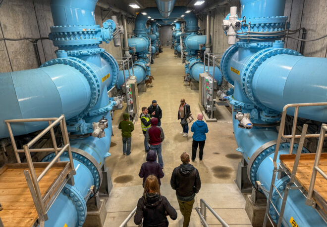 People walk through a water treatment facility lined with large blue pipes, valves, and filtration equipment.