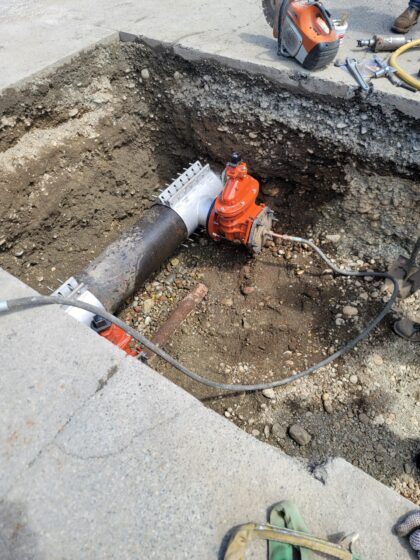 A construction site with an open excavation showing a dark water main pipe and an orange valve for a new water service installation, surrounded by tools and dirt.