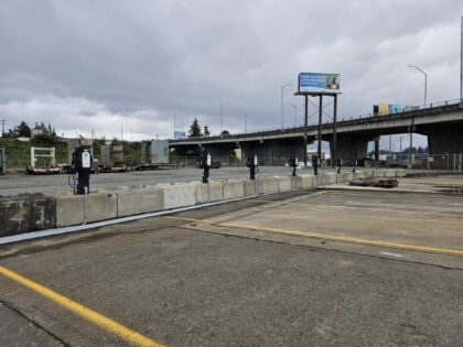 A row of temporary electric vehicle chargers under a concrete overpass, with shipping containers in the background and an overcast sky.