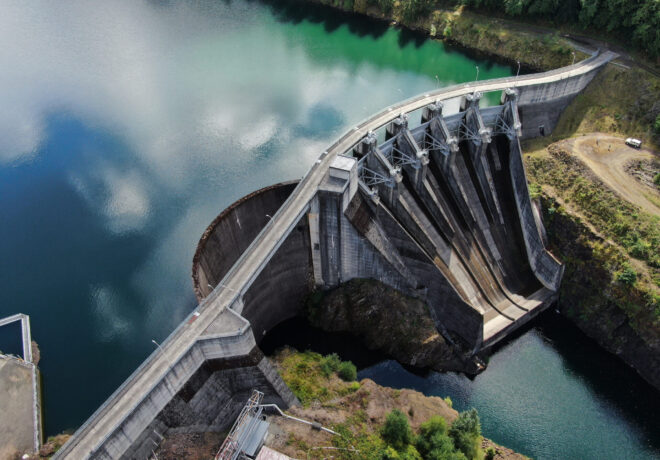 Aerial view of Mayfield Dam showing the curved concrete dam, spillway gates, reservoir, and river below, surrounded by forested hillsides.
