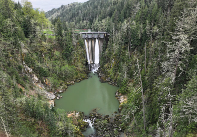 Aerial view of LaGrande Dam, spillway, and downstream river surrounded by evergreen forest.