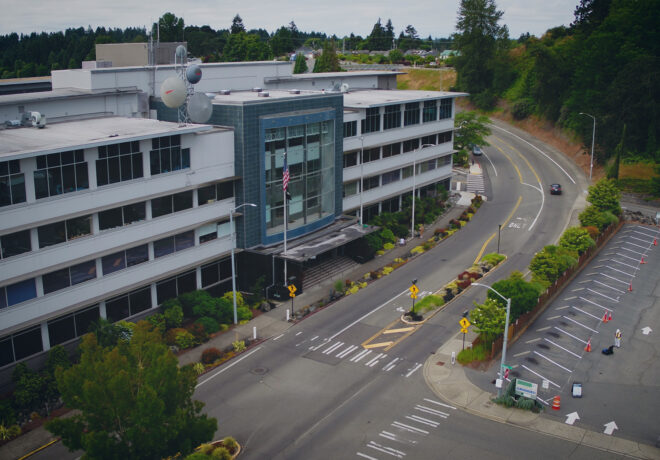Aerial view of a modern office building beside a curving road, landscaped sidewalks, and a mostly empty parking lot surrounded by trees.