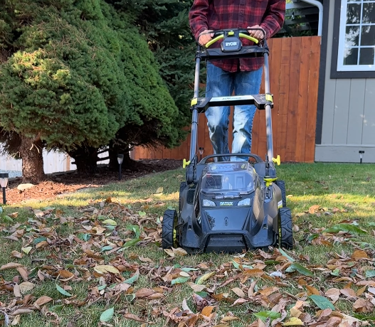 Person operating a black and green lawn mower on a leaf-covered lawn beside a house.