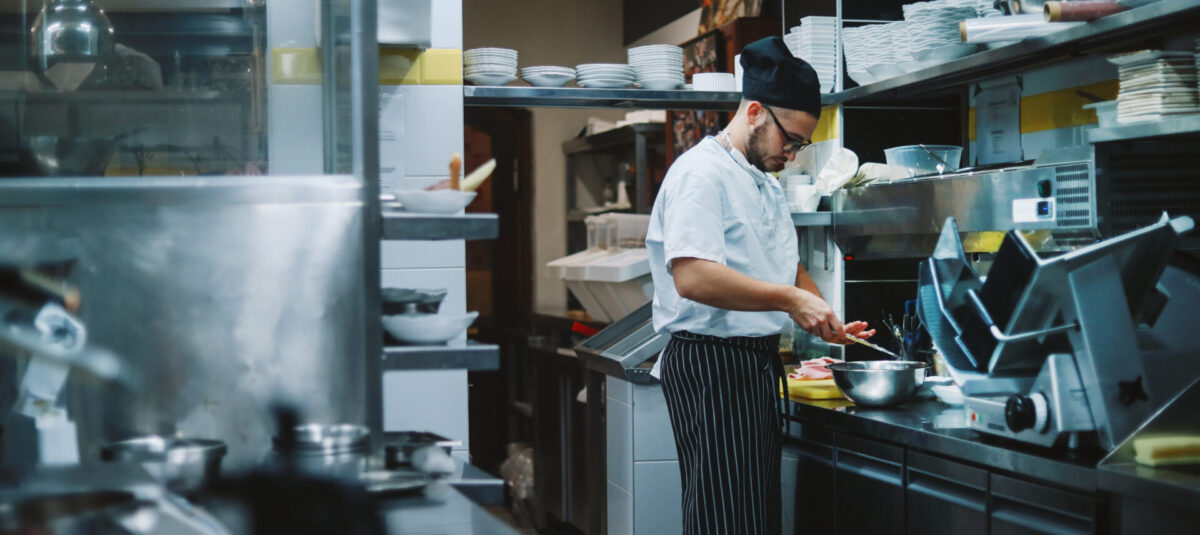 Chef preparing food in a busy commercial kitchen surrounded by stainless-steel equipment and stacks of white dishes.