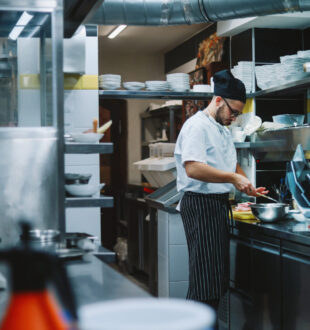 Chef preparing food in a busy commercial kitchen surrounded by stainless-steel equipment and stacks of white dishes.