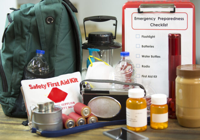 Various emergency preparedness supplies, including a green backpack, first aid kit, radio, water bottles, and a checklist, arranged on a table.