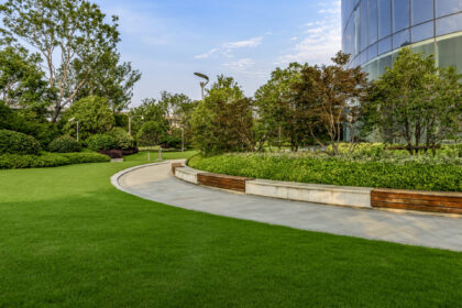 Landscaped courtyard with a curved walkway, wooden benches, lush greenery, and a modern glass building.