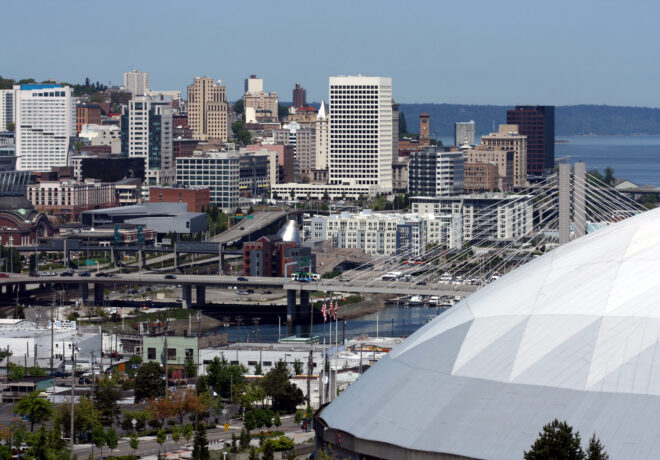 Panoramic view of a downtown waterfront city with high-rise buildings, elevated roadways, a stadium roof with cable supports, and a forested shoreline across the water.