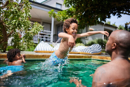 A child is leaping into a backyard pool toward an adult who is standing in the water, while another child swims nearby. Water splashes around them, and a house with a porch and striped lounge cushions is visible in the background.
