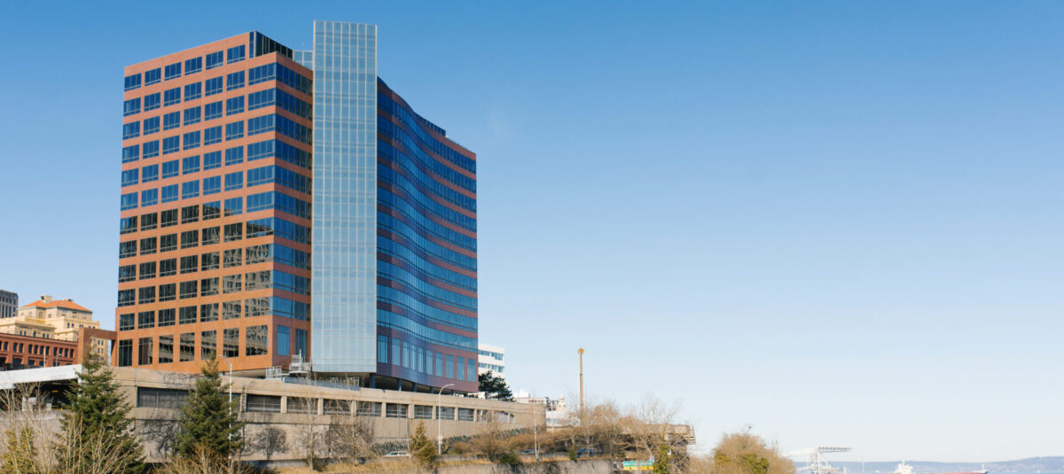 Modern glass-and-brick office buildings overlooking railway tracks, roads, trees, and a waterfront under a clear blue sky.