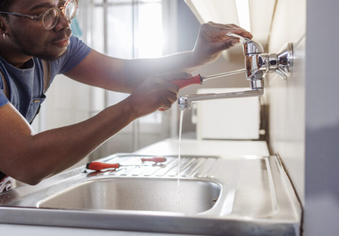 Person repairs a leaking kitchen faucet with a screwdriver while water runs into a stainless-steel sink.