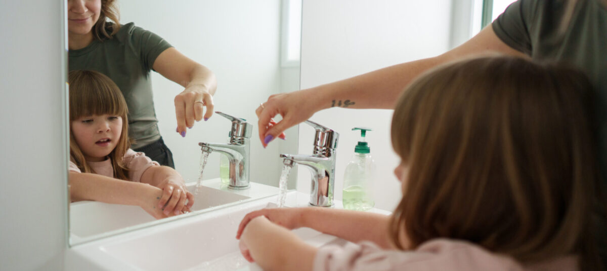 A woman helps a young girl wash her hands at a bathroom sink while they look in the mirror.