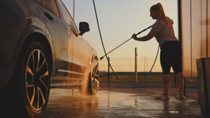 A person is spraying water onto the side of a car using a high‑pressure hose at an outdoor car wash. The car’s surface reflects warm sunset light, and water mist is visible around the wheels. The person stands on a wet concrete surface with a hose extended toward the vehicle, while a metal fence and open sky appear in the background.