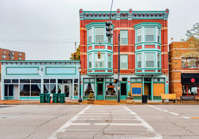A street view of three buildings, including a red brick structure with teal trim, a light blue single-story building, and a brick building with a black storefront. Trash bins are visible in the foreground.