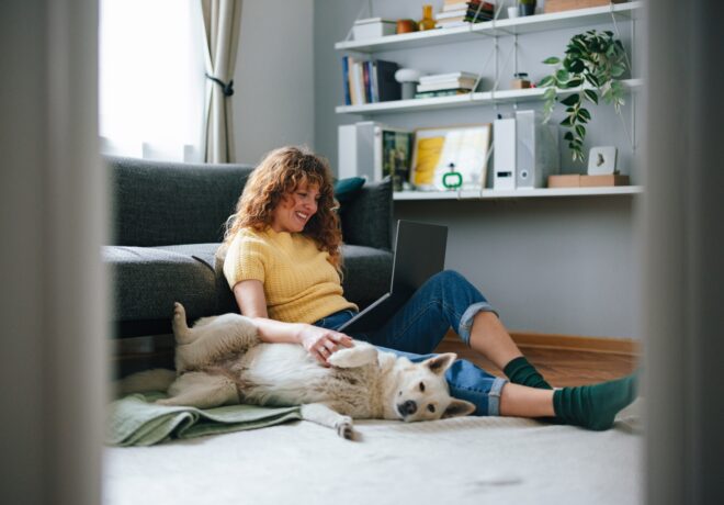 An individual sits on the floor next to a sofa with a laptop on their lap, gently resting a hand on a relaxed dog lying on its back. The room includes a bookshelf with plants, books, and household items in the background.