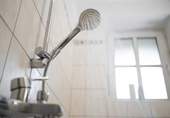 Chrome handheld showerhead spraying water in a tiled bathroom beside a bright window.