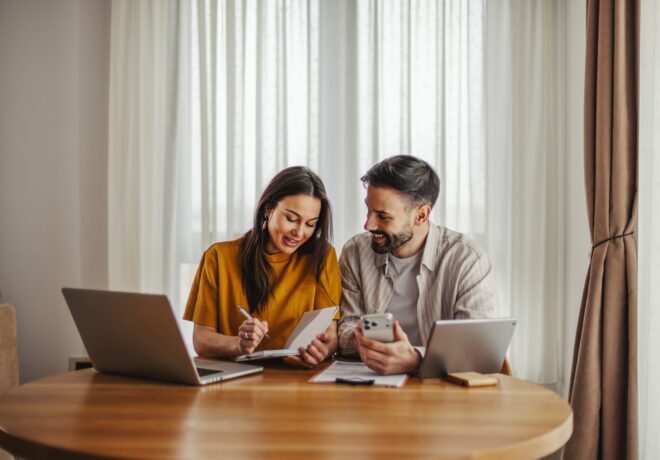 Young couple working together on their financial planning, reviewing documents and using digital devices like a laptop, smartphone, and tablet at home