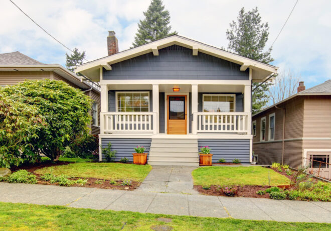 A blue residential house with white trim and a central porch, surrounded by green grass and sidewalks, features potted flowers by the entrance.