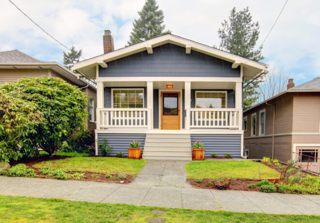 Single-story blue-gray house with a front porch, surrounded by a lush yard and a concrete pathway.