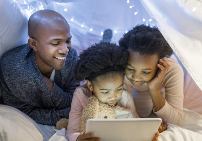 Smiling father looking at daughters using digital tablet in illuminated tent. Family is spending leisure time at home. They are wearing pajamas.