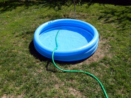 A small round blue inflatable kiddie pool sits on a grassy lawn, partially filled with water. A green garden hose lies across the grass and into the pool, suggesting it is being filled. The surrounding yard has patches of sunlight and a few bare spots in the grass.