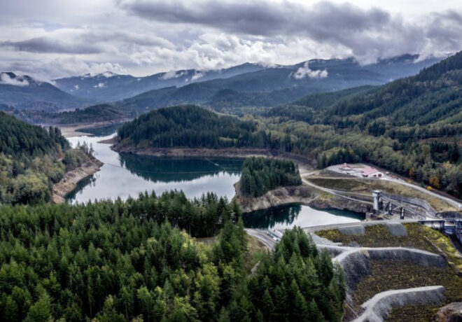 Aerial view of a forested mountain watershed with a reservoir, dam infrastructure, and access roads beneath cloudy mountain ridges.