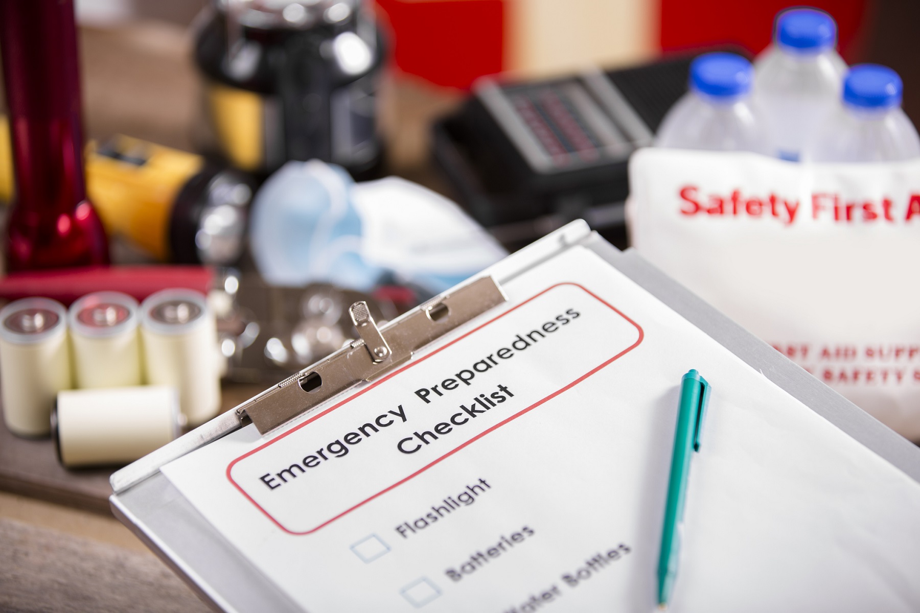 An image an Emergency preparedness checklist and pile of supplies to be used in case of a natural disaster (hurricane, flood, etc.): flashlight, batteries, water bottles, first aid kit, lantern, radio, mask. No people.