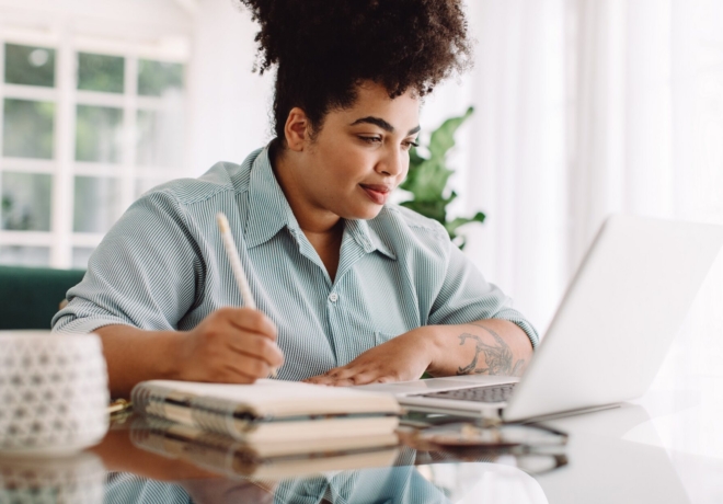Business woman working from home writing notes while looking at laptop. Confident woman sitting at desk using laptop and taking notes.
