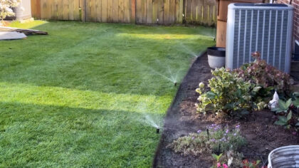 Backyard lawn being watered by sprinklers beside a flower bed, air-conditioning unit, and wooden fence.