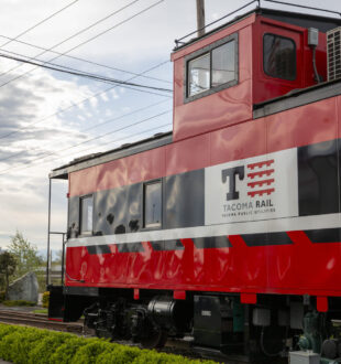 Red, white and black Tacoma Rail caboose in front of the Tacoma Rail facility. There are cloudy skies behind the caboose.