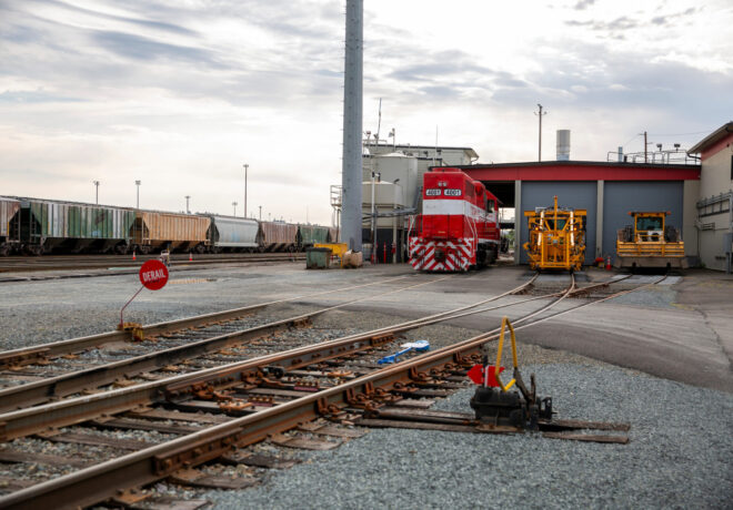 A railroad yard showing a red locomotive, various freight train cars, and a yellow maintenance vehicle. A red 
