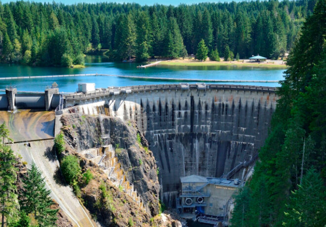 Drone view of Cushman Dam No. 2 with its curved concrete dam, reservoir, downstream channel, and surrounding evergreen forest.