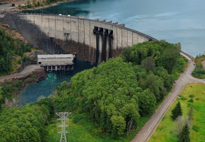 Aerial view of Mossyrock Dam on the Cowlitz River, showing the concrete arch dam, powerhouse, transmission substation and reservoir surrounded by forested hills.