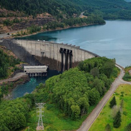 Aerial view of Mossyrock Dam on the Cowlitz River, showing the concrete arch dam, powerhouse, transmission substation and reservoir surrounded by forested hills.