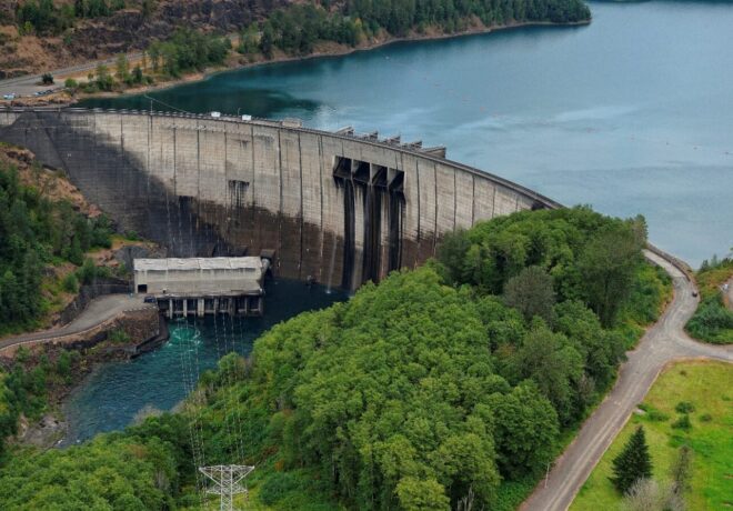 Aerial view of Mossyrock Dam on the Cowlitz River, showing the concrete arch dam, powerhouse, transmission substation and reservoir surrounded by forested hills.