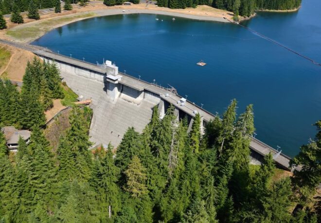 Drone view of Wynoochee Dam with the concrete dam spanning the reservoir, surrounded by evergreen forest and shoreline.