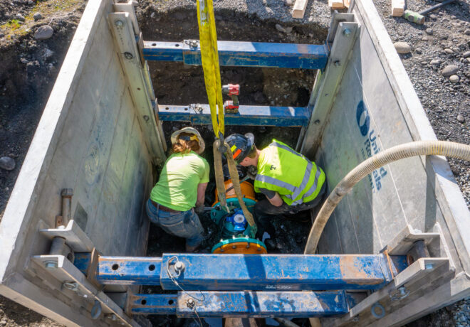 Two Tacoma Water workers in a trench are repairing a water main line, one in a neon green shirt and the other in a yellow vest, surrounded by concrete walls and tools.