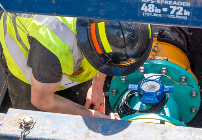 Worker in a reflective safety vest and hard hat handling cables and pipe machinery components.