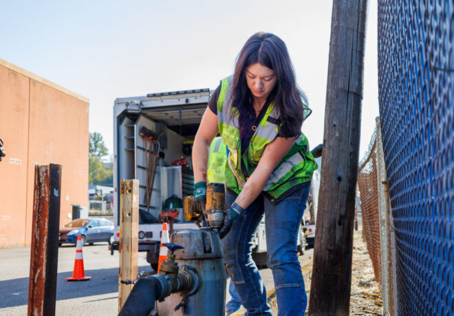 Worker in a reflective safety vest operates a heavy power tool on a metal pipe assembly beside a utility truck and chain-link fence.