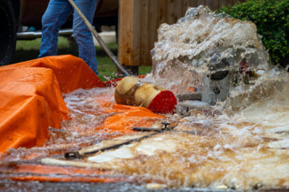 Fire hydrant flushing a large volume of water into an orange containment tarp, with a hose and worker visible in the background.