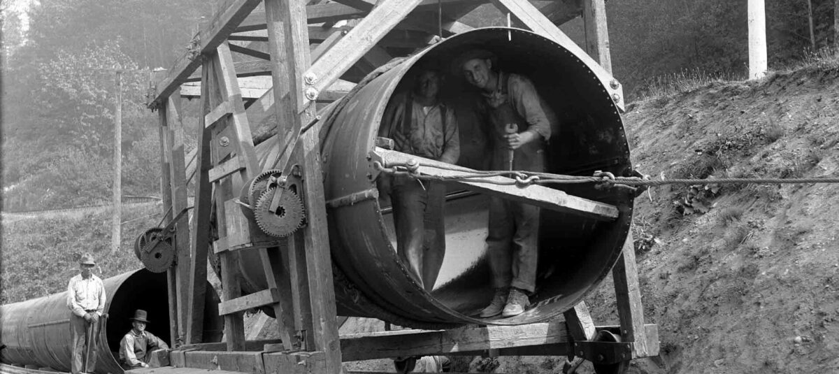 Workers construct a large water pipeline, with two men inside a pipe mounted on a wooden rail cart and others standing nearby.