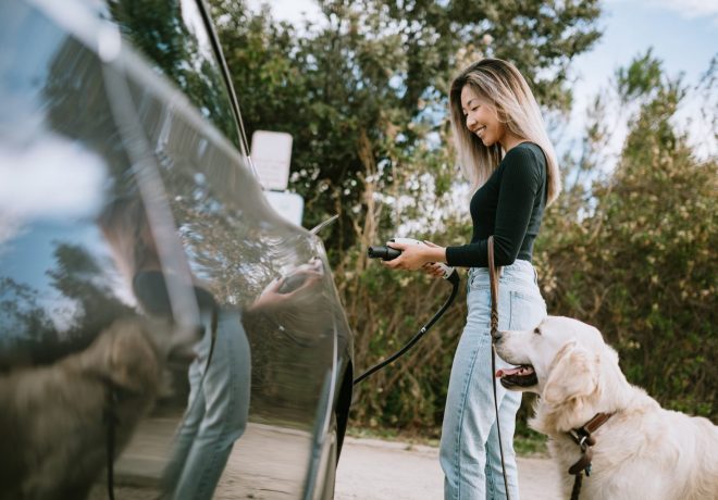 A woman and her dog stand next to an electric vehicle - the woman is plugging in a charger