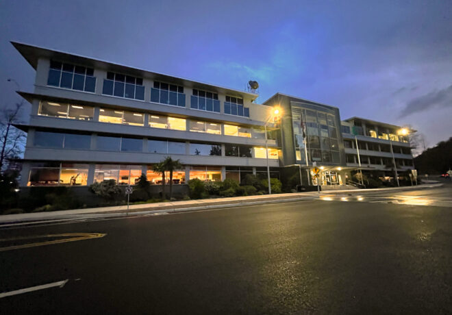 Modern glass-fronted building illuminated at dusk, viewed across a wet reflective street (TPU building).