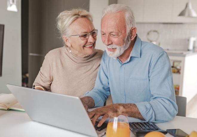 A senior couple is at home, they are sitting at the table and using a laptop