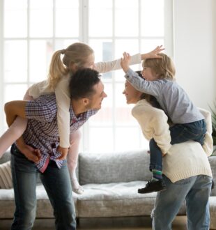 An image of a Happy young parents piggybacking son and daughter at home, smiling father and mother holding little kids on back spending time together, family having fun enjoying activity playing with children