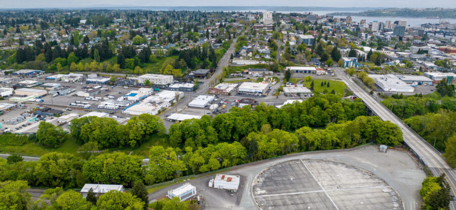 Aerial panorama of a tree-filled city with industrial buildings, roads, a large oval paved structure, and waterfront buildings under cloudy skies.