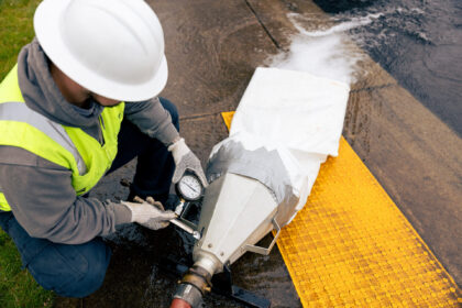 Worker in safety gear checks a pressure gauge on a large wrapped pipe assembly with exiting water beside yellow tactile paving.